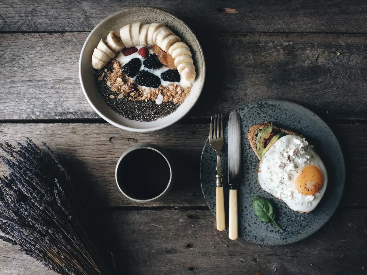 ชามข้าวโอ๊ตโรยกล้วยและผลไม้ วางข้างไข่ดาวและกาแฟบนโต๊ะไม้