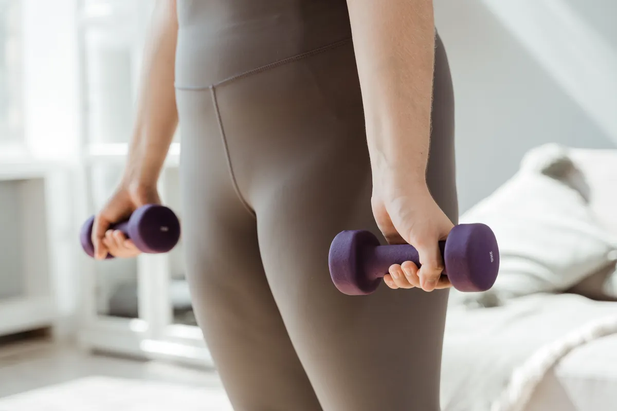 A woman in grey leggings holding a purple dumbbell in each hand in a room at home