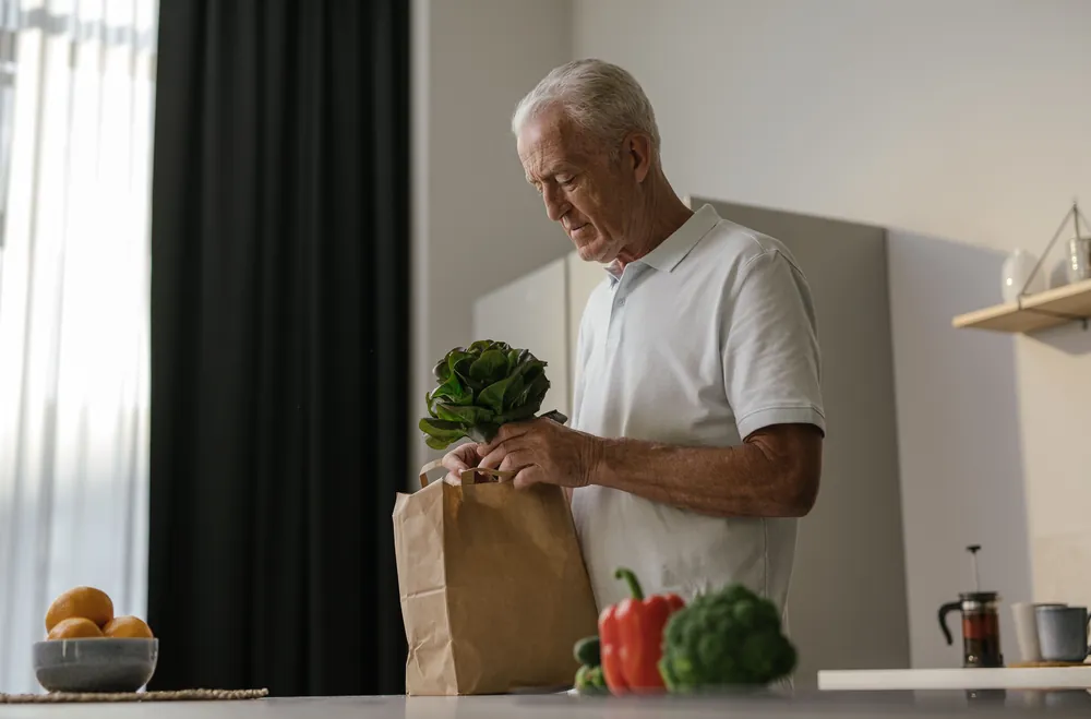 An older man taking leafy greens out of a paper bag in his kitchen